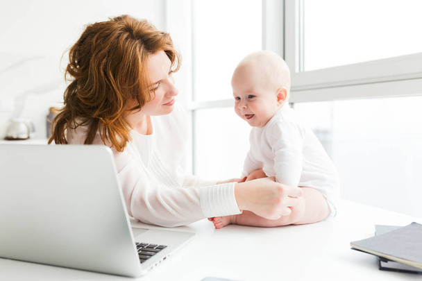 Portrait of beautiful mother sitting and dreamily looking at her happy little baby with laptop near isolated - Фото, изображение