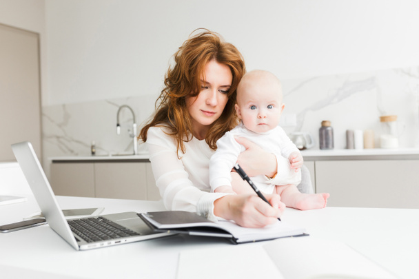 Portret van mooie zakenvrouw houden haar schattige kleine baby terwijl zitten aan de tafel en het maken van aantekeningen in notitieblok geïsoleerd - Foto, afbeelding