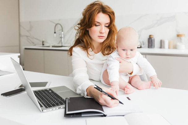 Close-up foto van zakenvrouw houden van haar kleine baby terwijl zitten aan de tafel en het maken van aantekeningen in notitieblok geïsoleerd - Foto, afbeelding