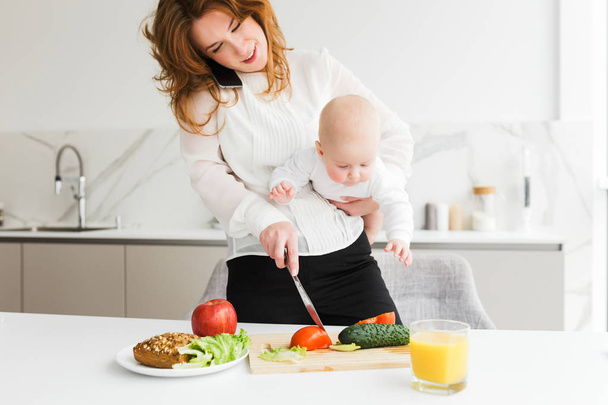 Portrait of pretty smiling mother standing and holding her little baby while talking on her cellphone and cooking on kitchen isolated - Photo, Image