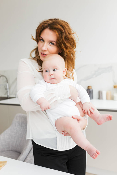 Porträt der schönen Mutter stehend und glücklich in die Kamera schauend, während sie ihr süßes kleines Baby isoliert in den Händen hält - Foto, Bild
