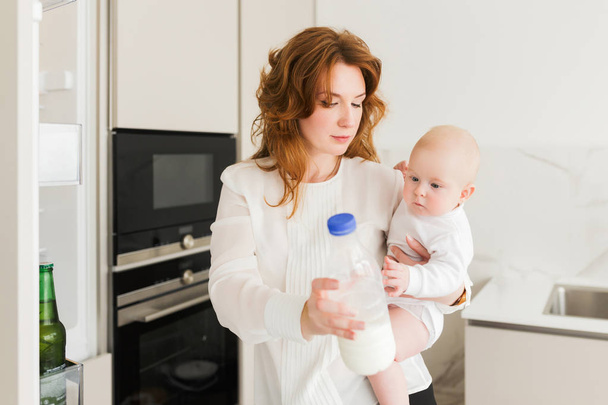 Πορτρέτο του στοχαστικός μητέρα στέκεται και κρατώντας το χαριτωμένο μικρό μωρό με μπουκάλι γάλα στο χέρι στην κουζίνα που απομονώνονται - Φωτογραφία, εικόνα