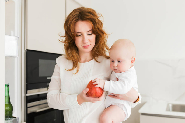 Portrait of beautiful smiling mother standing on kitchen and holding her cute little baby and big red apple in hands isolated - Photo, Image