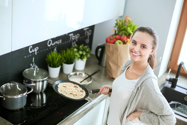 Young woman prepares pancakes in the kitchen while standing near the table. Woman in the kitchen. Cooking at kitchen. - Foto, Bild