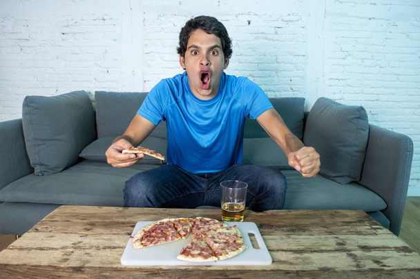 Young football fan celebrating a goal during watching football game on couch. Man watching soccer with beer and pizza - Foto, Imagen