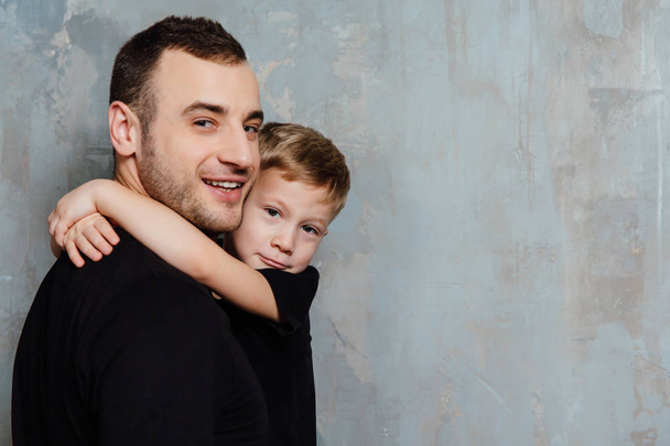 Vater und Sohn umarmen Sohn auf grauem Wandhintergrund. Mode Mann und Junge in schwarzen Kleidern. - Foto, Bild
