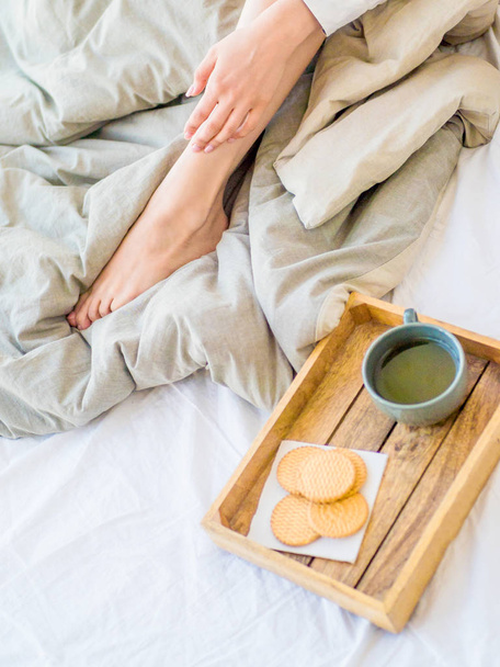 καφέ στο κρεβάτι. σχηματοποιημένα γυναικεία πόδια σε ζεστές κάλτσες, ξύλινο δίσκο για πρωινό στο κρεβάτι. Δύο φλιτζάνια καφέ και ζαχαρωτά. την έννοια του άνετου σπιτιού. πάνω όψη - Φωτογραφία, εικόνα