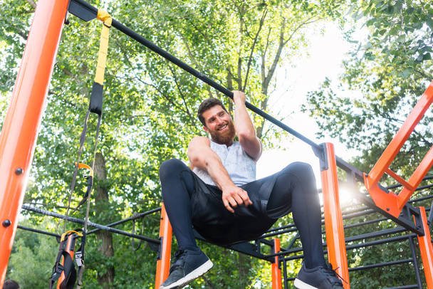 Powerful young man doing one-arm pull-ups while hanging on a bar in park - Photo, Image