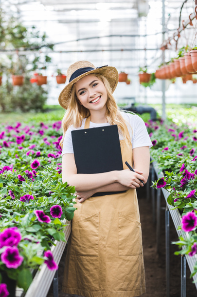Smiling female gardener holding clipboard by flowers in greenhouse - Photo, Image