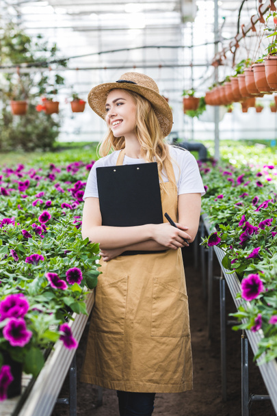 Attractive female gardener holding clipboard by flowers in greenhouse - Photo, Image