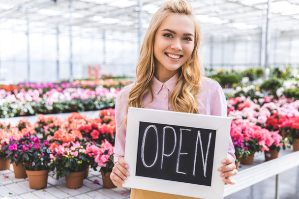 Attractive female gardener holding Open board by flowers in glasshouse - Photo, Image