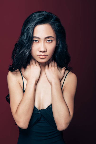 Young asian woman with hands on neck looking at camera. Sensual photo of girl in black dress on brown background - Foto, afbeelding