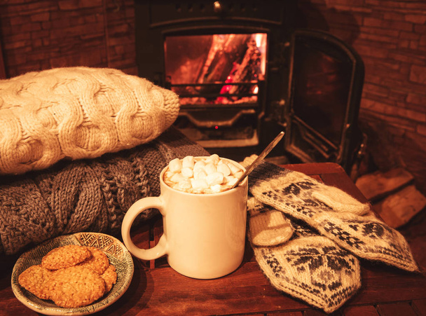 Стопка уютных вязаных свитеров, ручной работы варежки и чашки кофе, какао, горячего шоколада с зефиром на старом деревянном столе, рядом с горящим камином
 - Фото, изображение