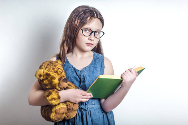 das Konzept von smart und lesen. Ein Mädchen im Schulalter mit Brille hält ein Buch, einen Bären und blickt in die Kamera. weißer Hintergrund.   - Foto, Bild