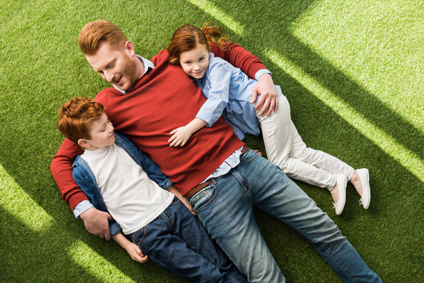 κάτοψη του χαρούμενος/η κοκκινομάλλης/α πατέρας με παιδιά ψέματα μαζί για την πράσινη χλόη - Φωτογραφία, εικόνα