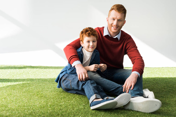 szczęśliwy ojciec i syn, siedząc na trawie i uśmiecha się do kamery na szary - Zdjęcie, obraz