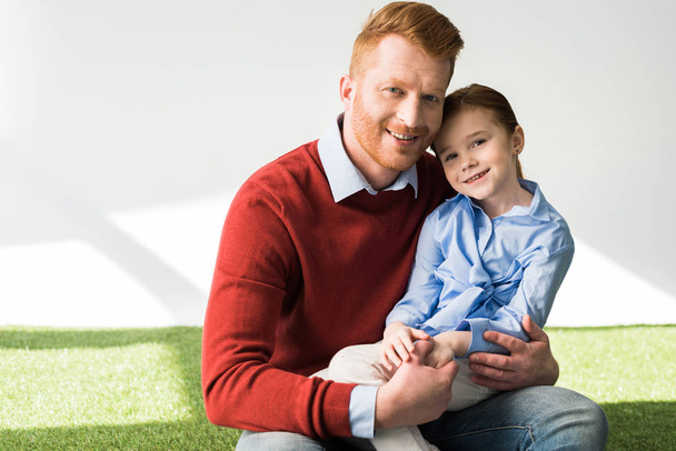 happy redhead father and daughter sitting on grass and smiling at camera on grey - Photo, Image