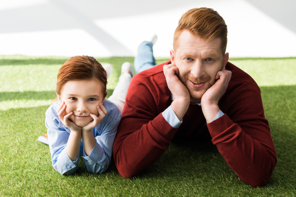 happy redhead father and daughter lying on grass and smiling at camera on grey  - Valokuva, kuva