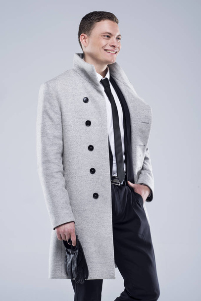 Jóképű fiatalember egy szürke kabátot és fekete öltöny áll egy szürke háttér - Fotó, kép