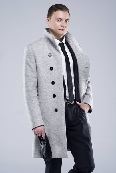 Handsome young man in a gray coat and black suit stands on a gray background - Foto, Imagen