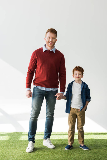full length view of happy redhead father and son holding hands and smiling at camera on grey  - 写真・画像