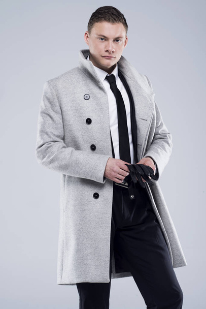 Handsome young man in a gray coat and black suit stands on a gray background - Фото, изображение