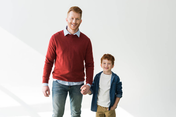 happy redhead father and son holding hands and smiling at camera on grey  - 写真・画像
