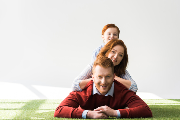 happy red haired family with one kid lying together on grass and smiling at camera on grey  - Фото, зображення