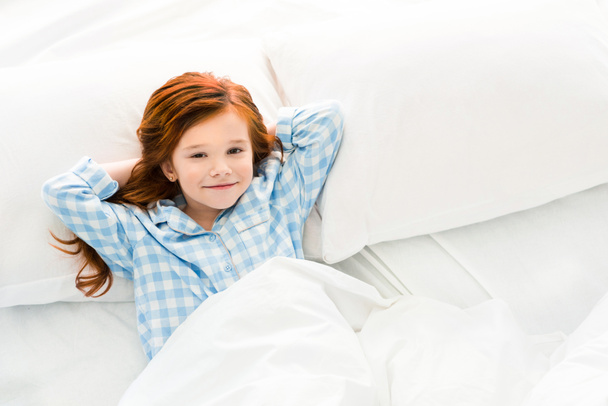 αξιολάτρευτο μικρό παιδί με τις πυτζάμες ξαπλωμένη στο κρεβάτι και να χαμογελά στη φωτογραφική μηχανή - Φωτογραφία, εικόνα