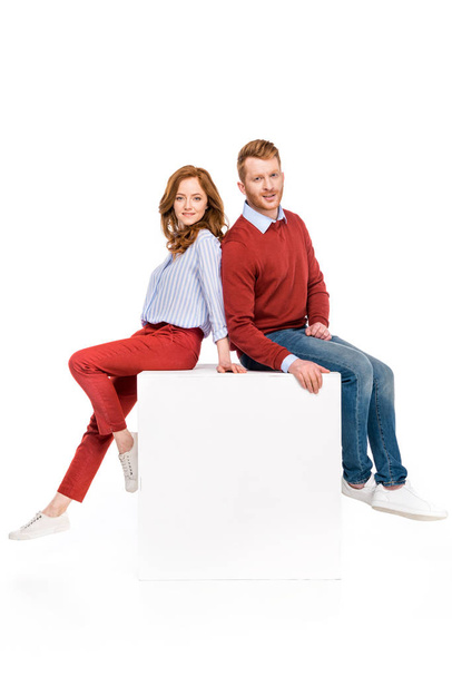 full length view of happy redhead couple sitting on white cube and smiling at camera isolated on white - Fotoğraf, Görsel
