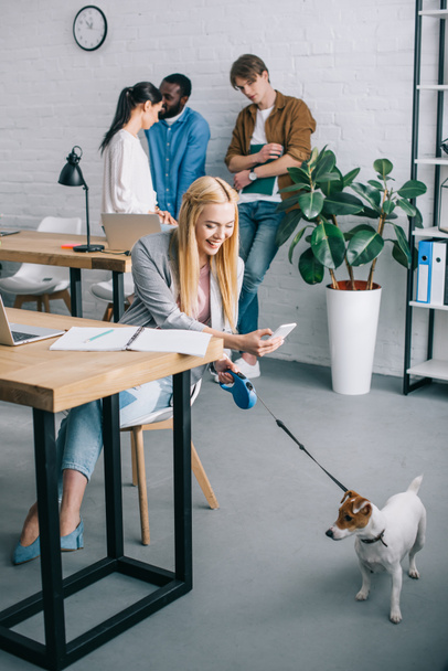 χαμογελώντας επιχειρηματίας λαμβάνοντας εικόνα από το σκυλί στο λουρί και συνεργάτες έχοντας συνάντηση πίσω από το σύγχρονο γραφείο  - Φωτογραφία, εικόνα