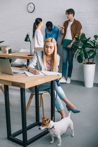 smiling businesswoman writing inn textbook and holding jack russell terrier on leash and coworkers having meeting behind in modern office  - Foto, imagen