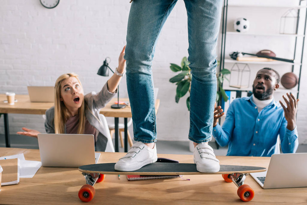 Oříznout obrázek podnikatel na longboard na stůl a šokován kolegy v moderní kanceláři   - Fotografie, Obrázek