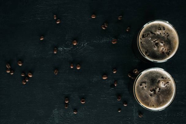 bovenaanzicht van glazen koude koffie met ijs op donkere oppervlakte met geroosterde koffiebonen rond - Foto, afbeelding