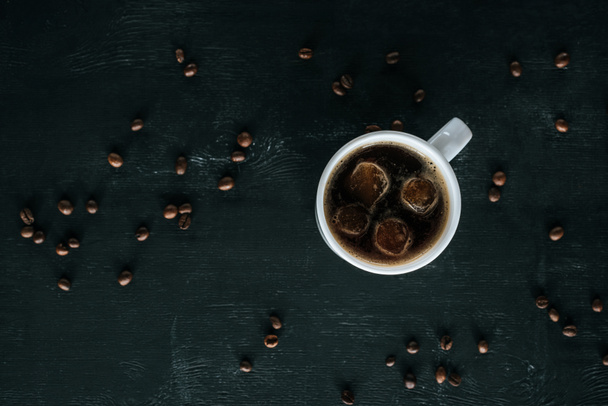 top view of mug of cold iced coffee on dark tabletop with roasted coffee beans around - Фото, изображение