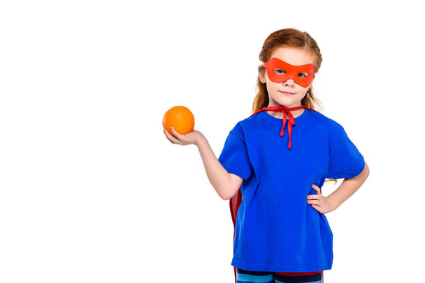 σούπερ παιδί στην μάσκα και μανδύα, κρατώντας πορτοκαλί και να χαμογελά στη φωτογραφική μηχανή που απομονώνονται σε λευκό  - Φωτογραφία, εικόνα