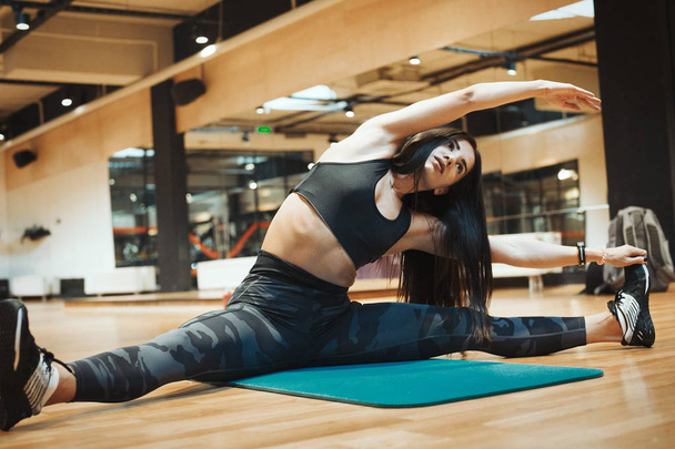 Young attractive brunette girl stretching and relaxing in asana pose at gym on the blue yoga mat after workout. Blurred background and young woman is on focus and foreground. Female do the splits - Photo, image