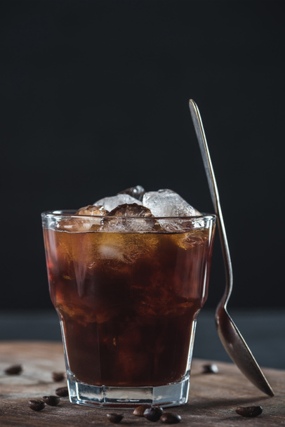 zblízka pohled na sklenici studené uvařené kávy se lžičkou na dřevěném prkénku na tmavé pozadí - Fotografie, Obrázek