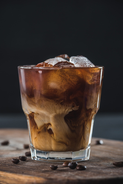 close up view of glass of cold brewed coffee with roasted coffee beans on wooden cutting board on dark backdrop - Valokuva, kuva