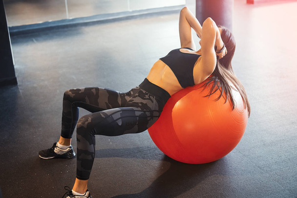 Young attractive brunette girl practicing workout and crossfit training and stretching on orange fitball. Blurred background and young woman is on focus and on foreground. - Fotografie, Obrázek