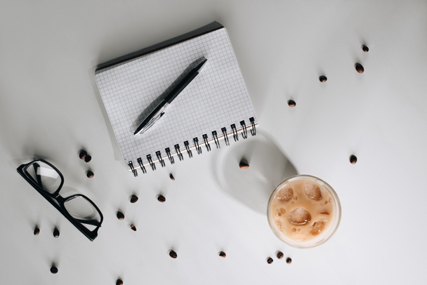 flat lay with glass of cold iced coffee, roasted coffee beans, eyeglasses, empty notebook and pen on white surface - Foto, imagen