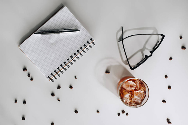 επίπεδη lay με ποτήρι κρύο παγωμένο καφέ, φρυγμένους κόκκους καφέ, γυαλιά, κενό σημειωματάριο και στυλό πάνω σε λευκή επιφάνεια - Φωτογραφία, εικόνα