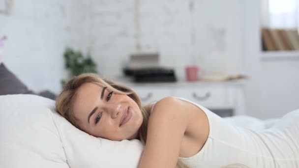 Pretty oung woman lying in the bed at the white pillow. Smiling and looking at camera. - Filmmaterial, Video