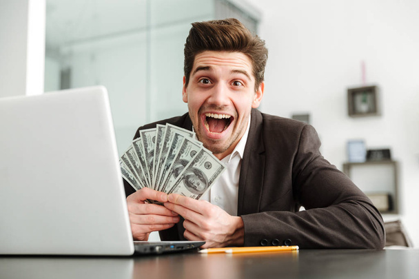 Ενθουσιασμένος νέος επιχειρηματίας δείχνει χρήματα που χρησιμοποιούν φορητό υπολογιστή. - Φωτογραφία, εικόνα