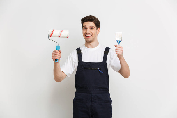 Happy male builder holding paint roll and brush - Fotó, kép