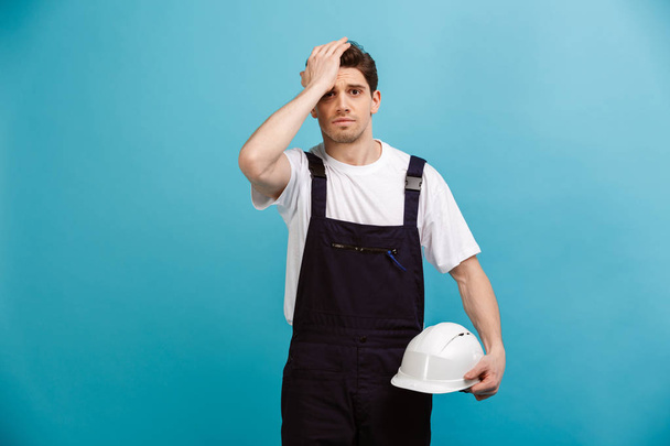 Worried male builder holding protective helmet while touching head - 写真・画像