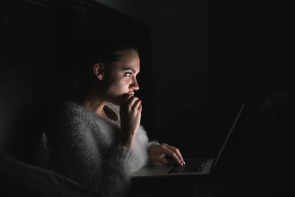 young girl sits in the dark at night, enthusiastically watches TV series on laptop - Фото, зображення