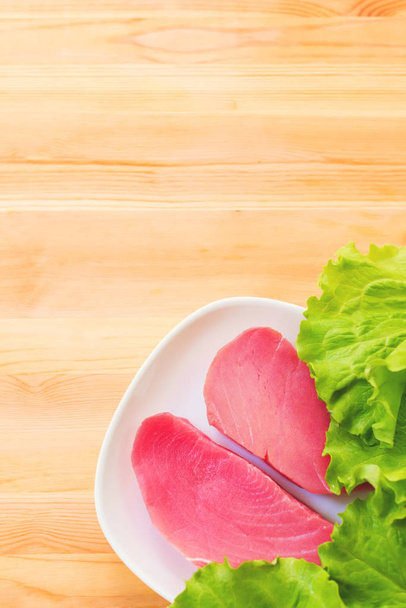 Two pieces of raw fresh tuna fillet on a plate with large leaves of a green salad on a wooden table. Delicious sea food easy. Greek cuisine - Foto, Imagem