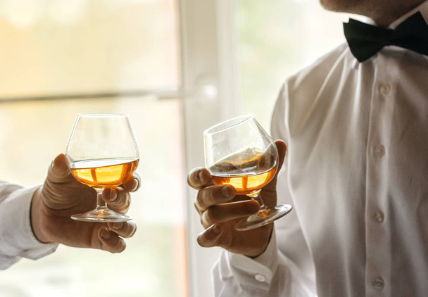 Dos vasos de whisky en las manos de hombres con camisas blancas y pajaritas. Tarde secular, brindis, amistad de hombres
. - Foto, imagen