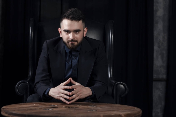Handsome attractive elegant classic man wearing trendy suit, with beard, sitting and having rest on luxury furniture in fashionable dark interior, A man in an expensive suit on the background of an ex - Photo, Image